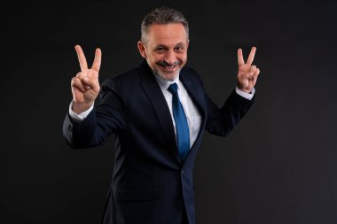 Takım elbiseli, kendine güvenen, gülümseyen ve iki elini de barış ya da zafer işareti olarak kaldıran, bir stüdyo çekiminde neşe, başarı ve pozitifliği ifade eden olgun bir adam..