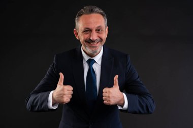 Resmi takım elbiseli, kendine güvenen, gülümseyen ve siyah arkaplandaki bir stüdyoda iki parmağını gösteren olgun bir adam, başarı ve onayı simgeliyor..