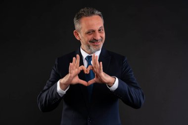 Takım elbiseli olgun bir işadamı elleriyle gülümseyip kalp şekli oluştururken profesyonel bir ortamda olumlu, özenli ve takdir dolu bir şekilde göz kırpıyor..