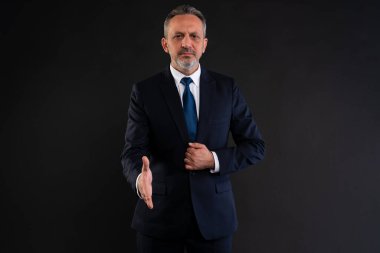 Siyah arkaplana karşı elini uzatan, anlaşmayı, ortaklığı ya da profesyonel selamlaşmayı sembolize eden koyu takım elbiseli, kendinden emin bir işadamı..
