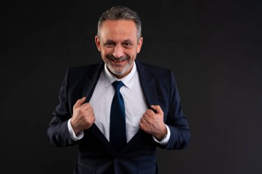 Takım elbiseli olgun bir işadamı, güvenle gülümsüyor ve siyah bir arkaplana karşı neşeli ve karizmatik bir pozla ceketini düzeltiyor..
