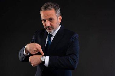 Resmi kıyafetli olgun bir adam, kendine güvenen ve kendine güvenen bir ifadeyle saatini işaret ederek, zamanın farkındalığını veya aciliyetini vurguluyor..