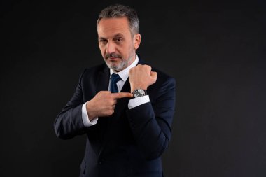 İş elbisesi giymiş olgun bir adam, ciddi bir ifadeyle zamanın önemini ya da aciliyetini gösteren kol saatine güvenle işaret ediyor..