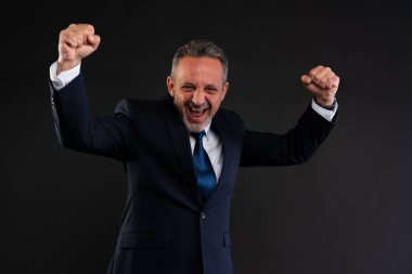 Takım elbiseli olgun bir adam kollarını kaldırarak kutluyor. Karanlık arka plan önünde sevinç ve heyecan ifade ediyor..