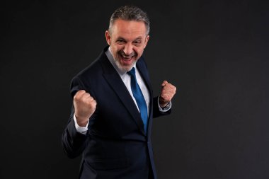 Orta yaşlı, takım elbiseli, yumruklarını sıkmış, geniş gülüşlü, stüdyo ortamında heyecanını ve zaferini ifade eden bir adam..