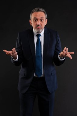 Orta yaşlı, takım elbiseli, kafası karışmış, omuzlarını siyah arka planda silkeleyen bir adam. Belirsizlik, kafa karışıklığı ya da cevap eksikliği kavramı.