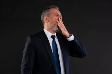 Resmi takım elbiseli orta yaşlı bir adam ağzını kapatıyor, yorgunluk ve bıkkınlık gösteriyor. Siyah arkaplanlı stüdyo görüntüsü.
