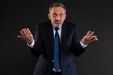 Takım elbiseli ve kravatlı olgun bir adam ellerini kaldırarak omuz silkiyor. Kafa karışıklığı ya da belirsizliği gösteriyor. Stüdyoda siyah arka planda çekilmiş..