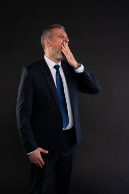 Takım elbiseli, kravatlı, esneyen ve ağzını eliyle kapatan yorgun bir işadamının portresi. Siyah arkaplanda stüdyo çekimi