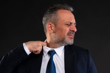 Takım elbiseli orta yaşlı bir işadamı gömleğinin yakasını rahatsızlıkla çekiyor. Sıcaklık ya da stres belirtileri gösteriyor. Siyah arkaplanda yakın çekim stüdyosu.