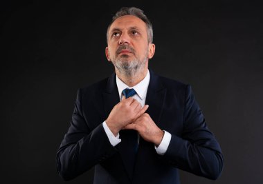 Resmi takım elbiseli orta yaşlı işadamı, siyah arka planda ciddi, yansıtıcı bir ifadeyle yukarı bakarken kravatını düzeltiyor..