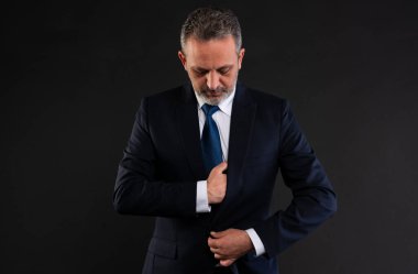 Olgun iş adamı resmi kıyafetini düzeltiyor, profesyonel bir stüdyo ortamında önünü iliklemeye odaklanıyor..