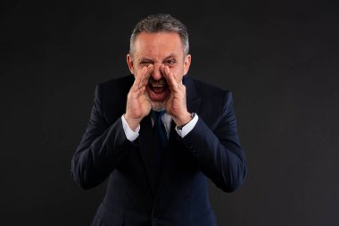 Takım elbiseli orta yaşlı bir adam elleri kolları bağlı bağırıyor, karanlık bir arka plana karşı hayal kırıklığı ya da aciliyet gösteriyor..