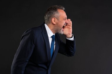 Orta yaşlı, donanma kıyafetli bir adam, eli ağzında, siyah bir arka plana karşı, yandan bağırıyor, aciliyet ya da öfke gösteriyor..