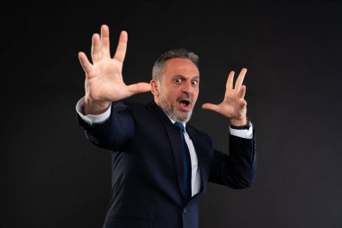 Takım elbiseli, şaşkın ve savunmacı görünen olgun bir adam. İki eli de önünde, karanlık bir geçmişe karşı ani bir duruma tepki veriyor gibi..