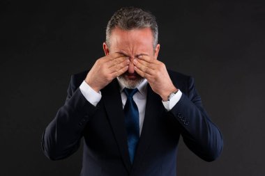 Resmi takım elbiseli olgun bir işadamı gözlerini iki eliyle ovuşturuyor, yorgunluğunu, hayal kırıklığını ya da stresi ifade ediyor, karanlık arka planda izole edilmiş..