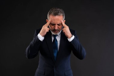 Takım elbiseli ve kravatlı olgun bir işadamı, parmaklarını şakaklarına bastırıyor. Stres, konsantrasyon ya da baş ağrısı ifade ediyor..