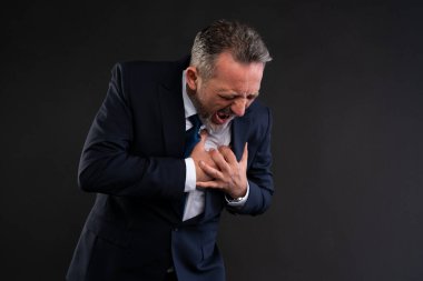 Takım elbiseli sıkıntılı olgun bir adam siyah arka planda bağırıyor ve göğsünü sıkıyor şiddetli kalp ağrısı ya da acil durum belirtileri gösteriyor..
