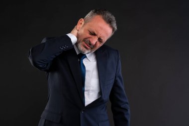 İş elbisesi giymiş orta yaşlı bir adam sıkıntılı bir şekilde boynunu tutuyor, ağrı ya da kasılma belirtileri gösteriyor, muhtemelen stres, kötü duruş ya da uzun çalışma saatleri yüzünden..