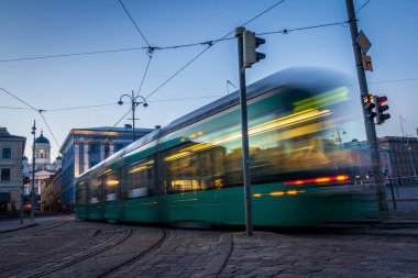 Şehirde toplu taşıma. İstasyondan ayrılan tramvay, trafik ışıkları, insanlar, sokak, evler, teller, raylar. Hareket ve ışıklar, uzun pozlama.