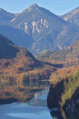 Sakin bir göl görkemli dağ zirvelerinin altındaki canlı sonbahar renklerini yansıtır. Dağlar yükselir, zirveleri batan güneşin sıcacık parıltısıyla yıkanır. Fussen, Almanya.