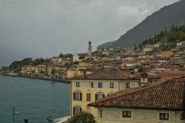 Bir dağ zeminine kurulmuş göl kenti manzarası. Terracotta çatıları ve rengarenk binalar rıhtım boyunca uzanıyor. Gölün sakin suları yukarıdaki bulutlu gökyüzünü yansıtır..
