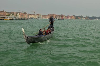 Venedik, İtalya - 15 Ekim 2024 Venedik 'in zümrüt yeşili gölünde bir gondol süzülüyor. Turistler bir gondolcunun rehberliğinde manzaranın tadını çıkarıyorlar. Sahilde tarihi yapılar görülebilir..