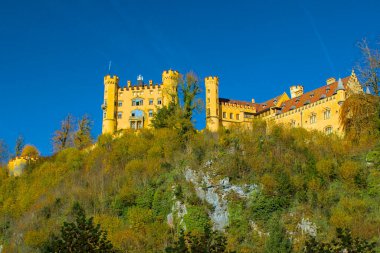 Görkemli Hohenschwangau Kalesi bir tepenin üstünde gururla durur. Sarı maskaralığı güneşli bir günün sıcacık parıltısıyla yıkanır. Bu tarihi Bavyera mücevheri çarpıcı bir dağ manzarasına bakıyor.. 
