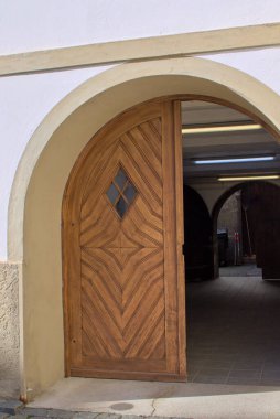 Karmaşık geometrik desenli büyük ahşap bir kapı hafifçe açık duruyor. Kemerli giriş, üzerinde floresan lambalar olan loş bir koridora çıkıyor ve derinlik hissi yaratıyor..