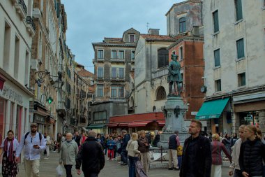 Venedik, İtalya - 16 Ekim 2024: Venedik, İtalya 'da hareketli bir cadde, şehrin eşsiz mimarisi ve atmosferini gözler önüne seriyor. İnsanlar dar geçitlerde dolaşıyorlar. Ortada dik duran bir heykel..