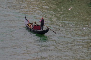 Venedik, İtalya - 16 Ekim 2024: Bir gondolcu dalgalı suyun ortasında yolcuları taşıyan Venedik lagününden bir gondol geçirir. Yalnız bir martı yükselir.