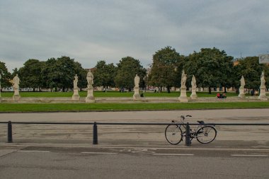 Padua, İtalya - 17 Ekim 2024: Klasik bir bisiklet tarihi Prato della Valle 'ye giden geniş bir yolda dinlenir. Ağaçlarla dolu parkı bulutlu bir gökyüzünün altında çerçeveleyen, yeşil alana uzanan heykeller..