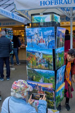 Schwangau, Almanya - 21 Ekim 2024: Turistler Schloss Neuschwanstein 'in kartpostallarına hayran. Bir kadın hevesle telefonuyla anın fotoğrafını çeker. Hediyelik eşya dükkanları ziyaretçileri çeker..