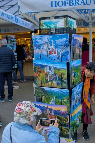 Schwangau, Almanya - 21 Ekim 2024: Turistler Schloss Neuschwanstein 'in kartpostallarına hayran. Bir kadın hevesle telefonuyla anın fotoğrafını çeker. Hediyelik eşya dükkanları ziyaretçileri çeker..