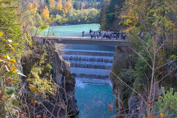 Fussen, Almanya - 21 Ekim 2024: Çarpıcı Lechfall 'a bakan bir köprü üzerinde bir grup insan duruyor. Titreşimli turkuaz su birden fazla kattan aşağı dökülür..
