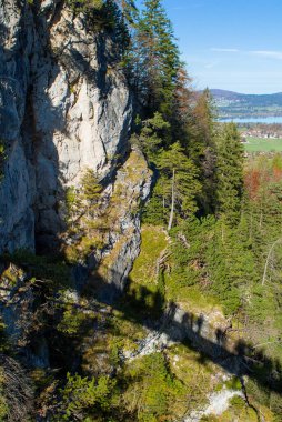 Almanya 'nın Schwangau kentindeki sık dağların yanında dramatik bir uçurum yükselir. Aşağıda, yeşil kayalar ve ağaçlar arasında uzanan gölgeli bir köprü var. Açık gökyüzü ve uzak tepeler bu çarpıcı manzarayı tamamlıyor..