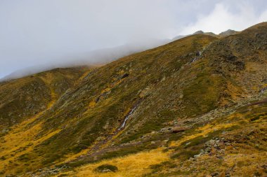 Altın otlar Timmelsjoch Geçidi 'nin engebeli yamaçlarını kaplar. Dar bir dere yavaşça kayalık yamaçtan aşağı akıyor. Alçak bulutlar ufuklarda saklanarak zirvede sürüklenir..