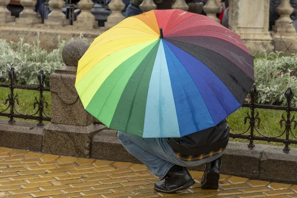 Crouched man with a rainbow colored umbrella during a rain shower.