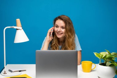 Dizüstü bilgisayar başında çalışırken telefonda konuşan kıvırcık saçlı bir kadın. Beyaz masada oturuyor ve gülümsüyor.