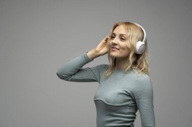Beyaz kulaklıklı sarışın kız stüdyoda gri arka planda müzik dinliyor.