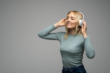 Mavi tişört ve gözlüklü, beyaz kulaklıklı, müzik dinleyen ve stüdyoda gri arka planda gülümseyen güzel sarışın kadın. Rahatlamak ve eğlenmek. Yaşam biçimi