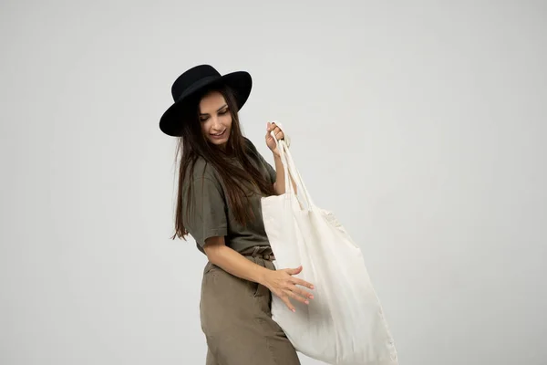 Brunette woman in green t-shirt and black hat holding cotton shopper bag with vegetables, products in white room. Eco friendly shopping bags. Zero waste, plastic free concept