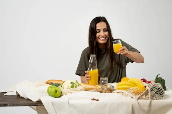 Woman in green t-shirt pouring a juice from a glass bottle in a glass over a table with mesh eco bag, healthy vegan vegetables, fruits, bread, snacks. Zero waste concept