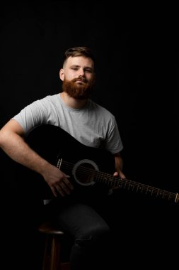 Young bearded guitarist in grey t-shirt holds a guitar and playing a song in a dark room