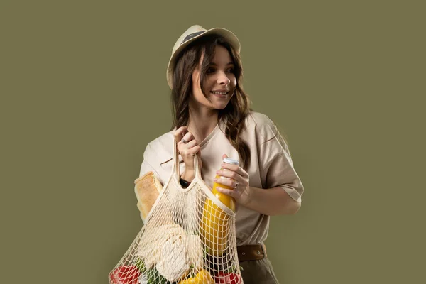 stock image Smiling young woman in light summer clothes with a mesh eco bag full of vegetables, greens watching in a camera on a green studio background. Sustainable lifestyle. Eco friendly concept. Zero waste