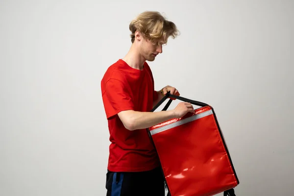 Young courier opening thermal bag on white background. Food delivery service. Delivery guy in a red t-shirt uniform work as courier and holds red thermal food backpack. Service concept
