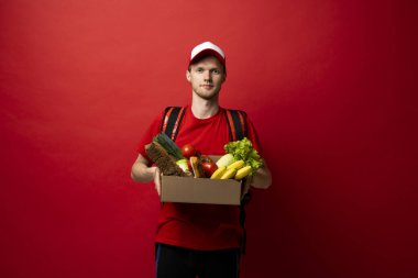 Teslimatçı, beyaz duvarın üzerinde yiyecek dolu bir kutu tutuyor. Çevrimiçi süpermarket. Yemek dağıtım servisi.