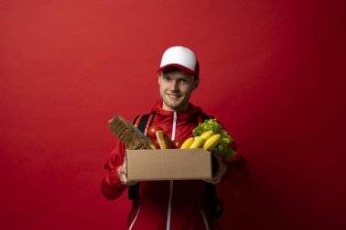 Kırmızı üniformalı, yiyecek, meyve ve sebze dolu kutular taşıyan işçi teslim edin. Postacı ve market teslimat hizmeti kavramı