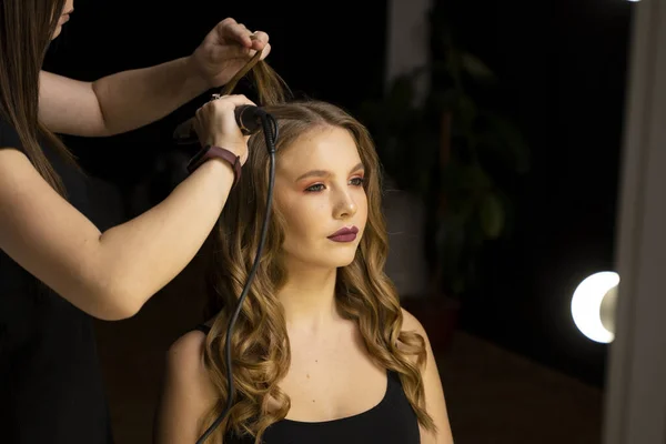 プロのスタイリストの美容師は カーリングアイロンで長い女性の髪をカールし 女性のクライアントのためのスタイリングを作ります ビューティーサロン ストックフォト