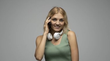 Gülümseyen, yeşil tişörtlü, beyaz kulaklıklı, cep telefonuyla konuşurken kameraya bakan mutlu, genç, sarışın kadın.